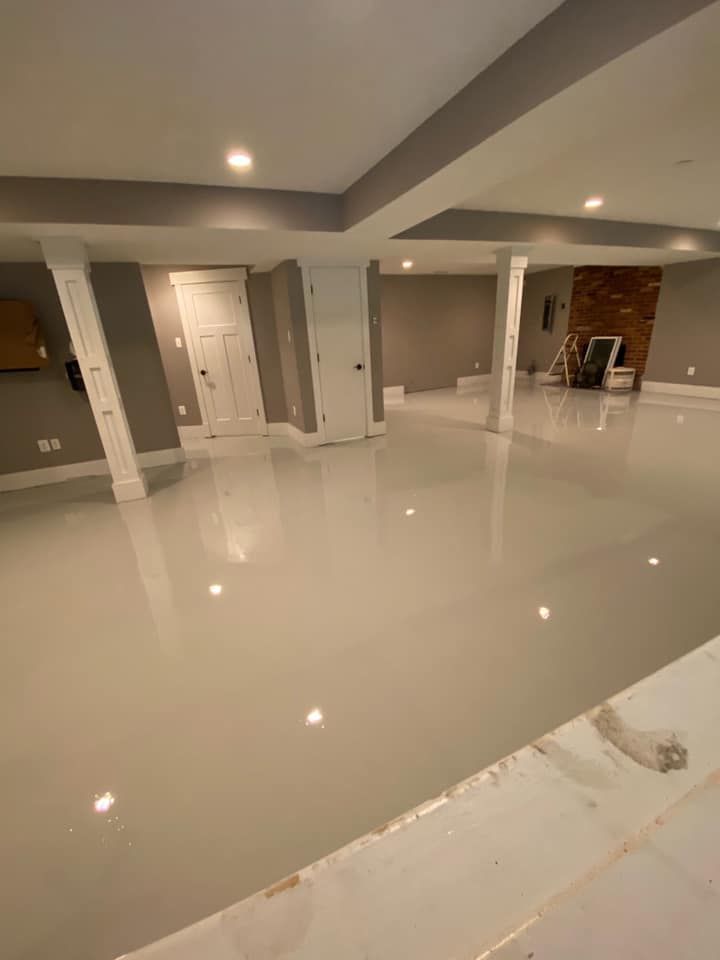 A bright, empty basement with a high-gloss, light gray floor, white structural columns, gray walls, and two white doors.