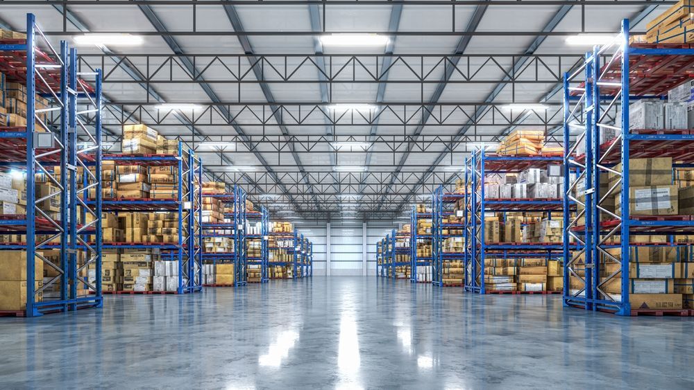 Wide aisle of a large, modern warehouse with blue metal shelving units filled with various boxes on both sides.
