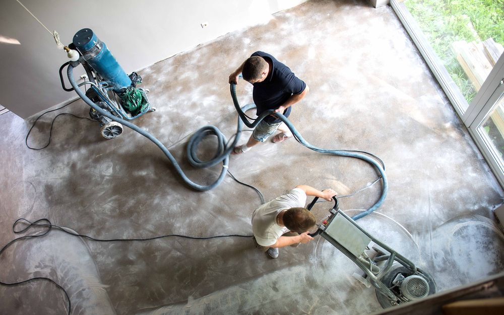 Two people use a floor sanding machine and vacuum system to smooth a concrete surface in an unfinished room.