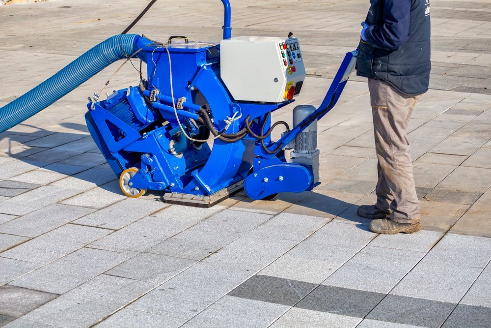 A person operates a blue portable surface shot blaster on stone paving tiles, connected to a long blue suction hose.