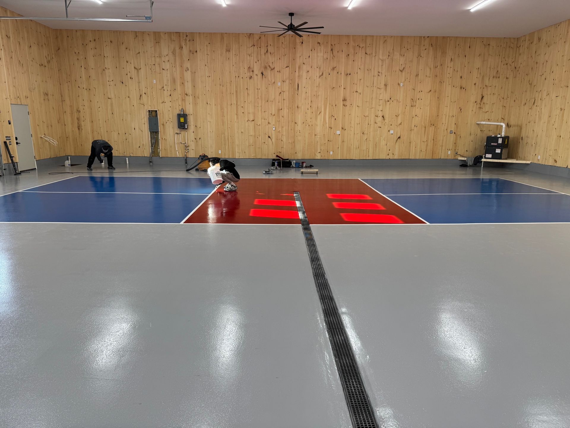 People working on a shiny garage floor with painted blue, red, and grey sections against wood-paneled walls.