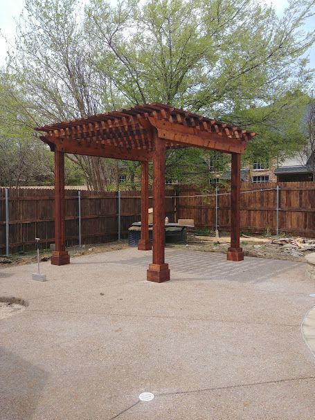 Wooden pergola on a concrete patio with a wooden fence backdrop and a tree.