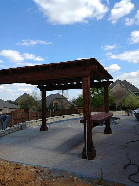 Wooden pergola over a concrete patio with a pool, brown stain, and a sunny sky.