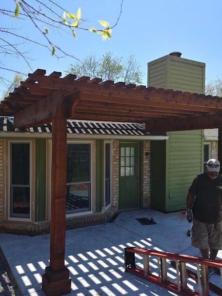 Pergola over patio in front of a house with green walls, a worker stands nearby.
