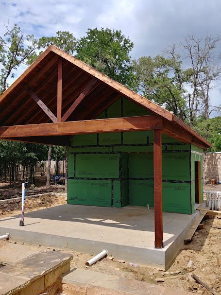 Building exterior under construction with green sheathing and a wood-beamed roof. Concrete patio and surrounding dirt.