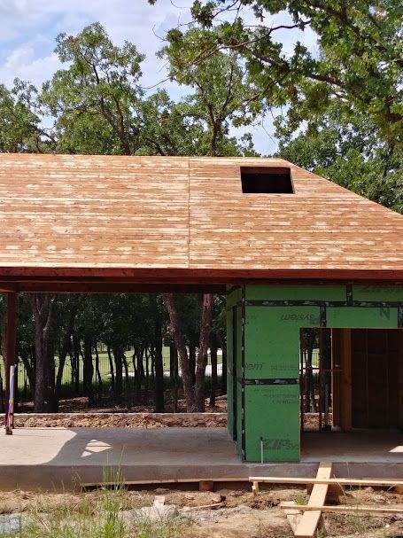 Construction site, building's roof with sheathing installed, green walls, and trees in the background.