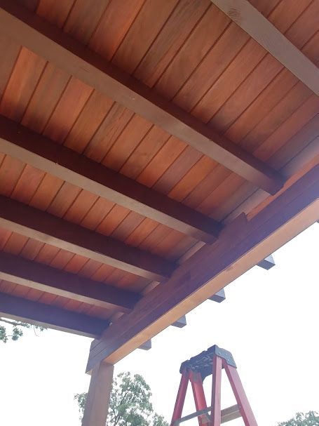 Wooden pergola ceiling with reddish-brown planks and dark beams; red ladder in the foreground.