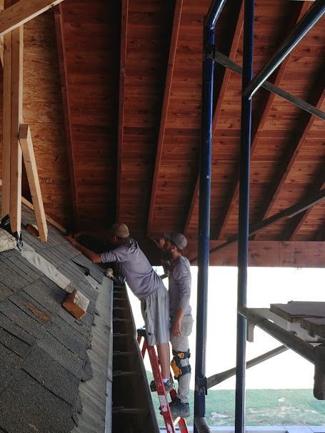 Two men on a ladder are repairing a roof. One works on the shingles, the other on a support beam.