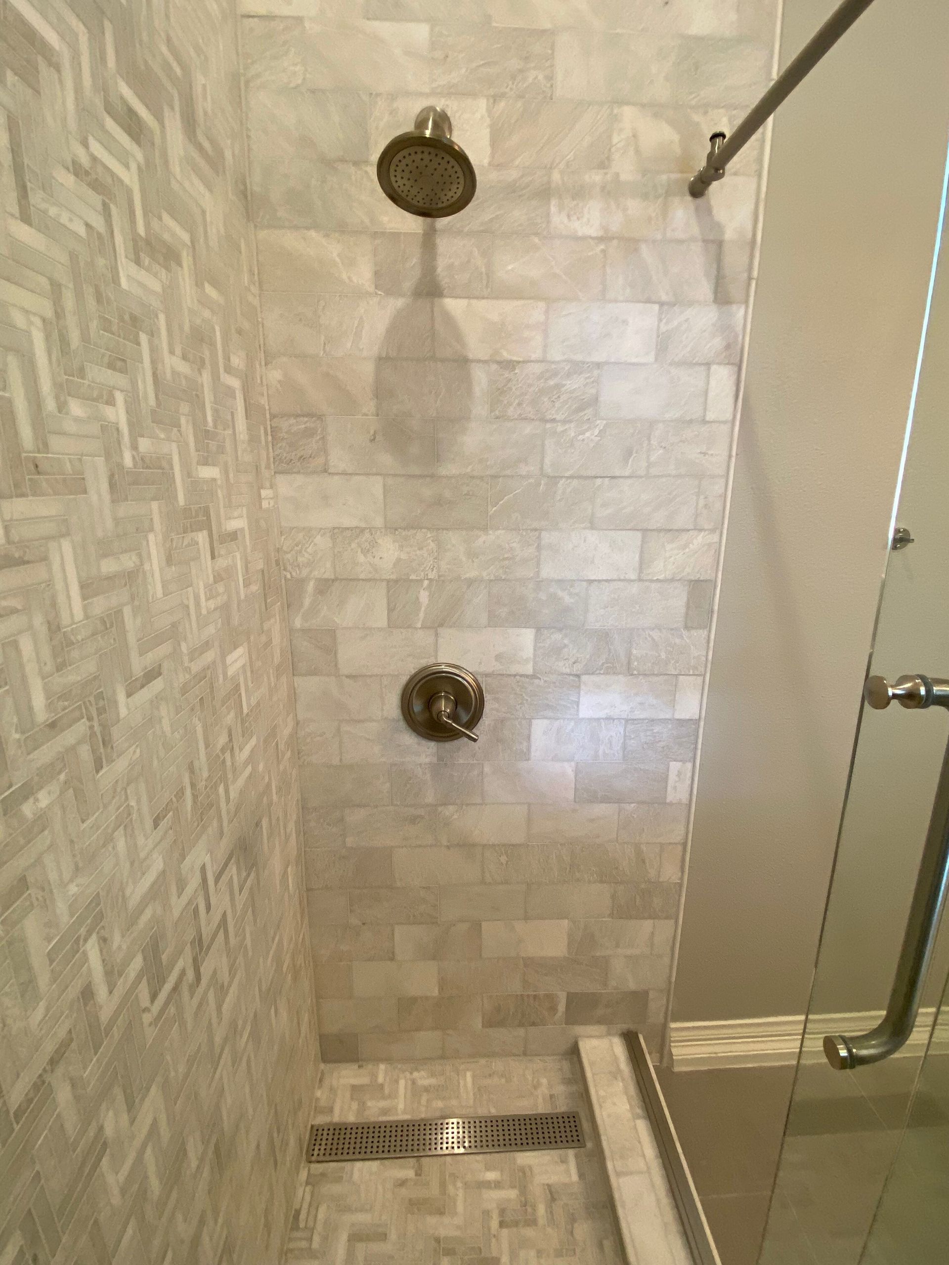 Shower with white and gray tile, a bronze showerhead, and a glass door.