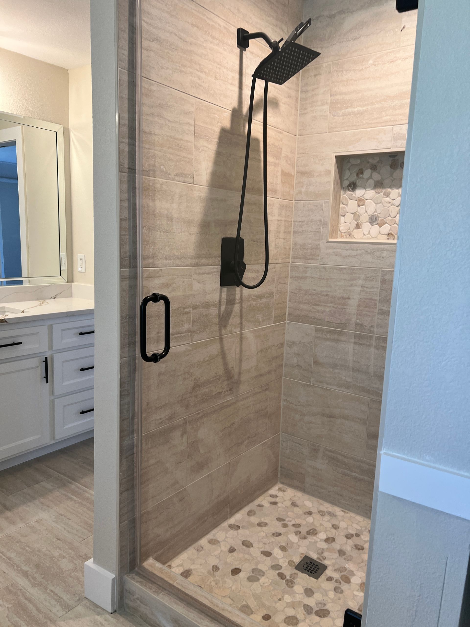 Bathroom with a glass shower, beige stone tiles, and black fixtures.