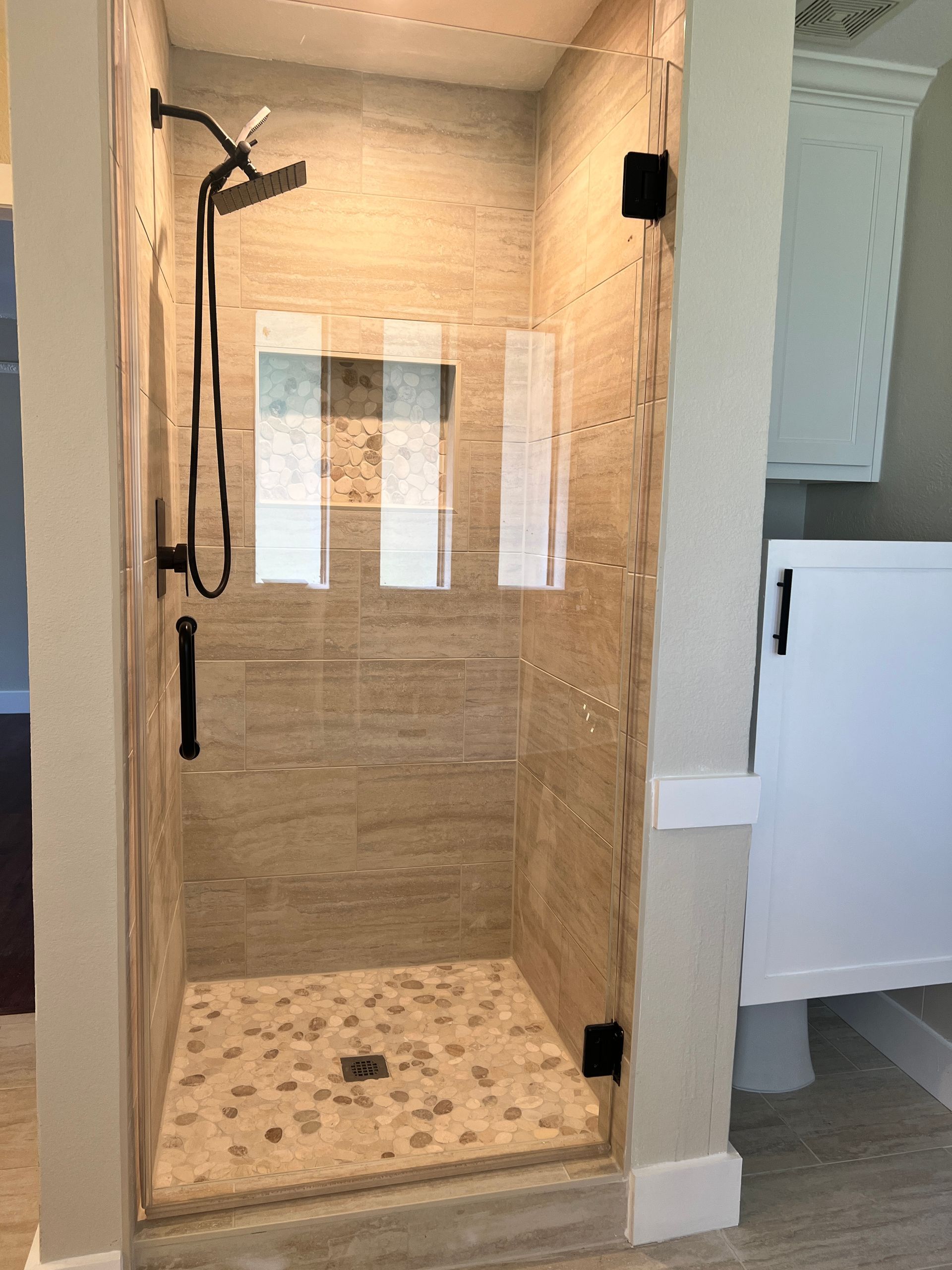 Modern shower with pebble floor and glass door, black fixtures, neutral tile, and white cabinet.
