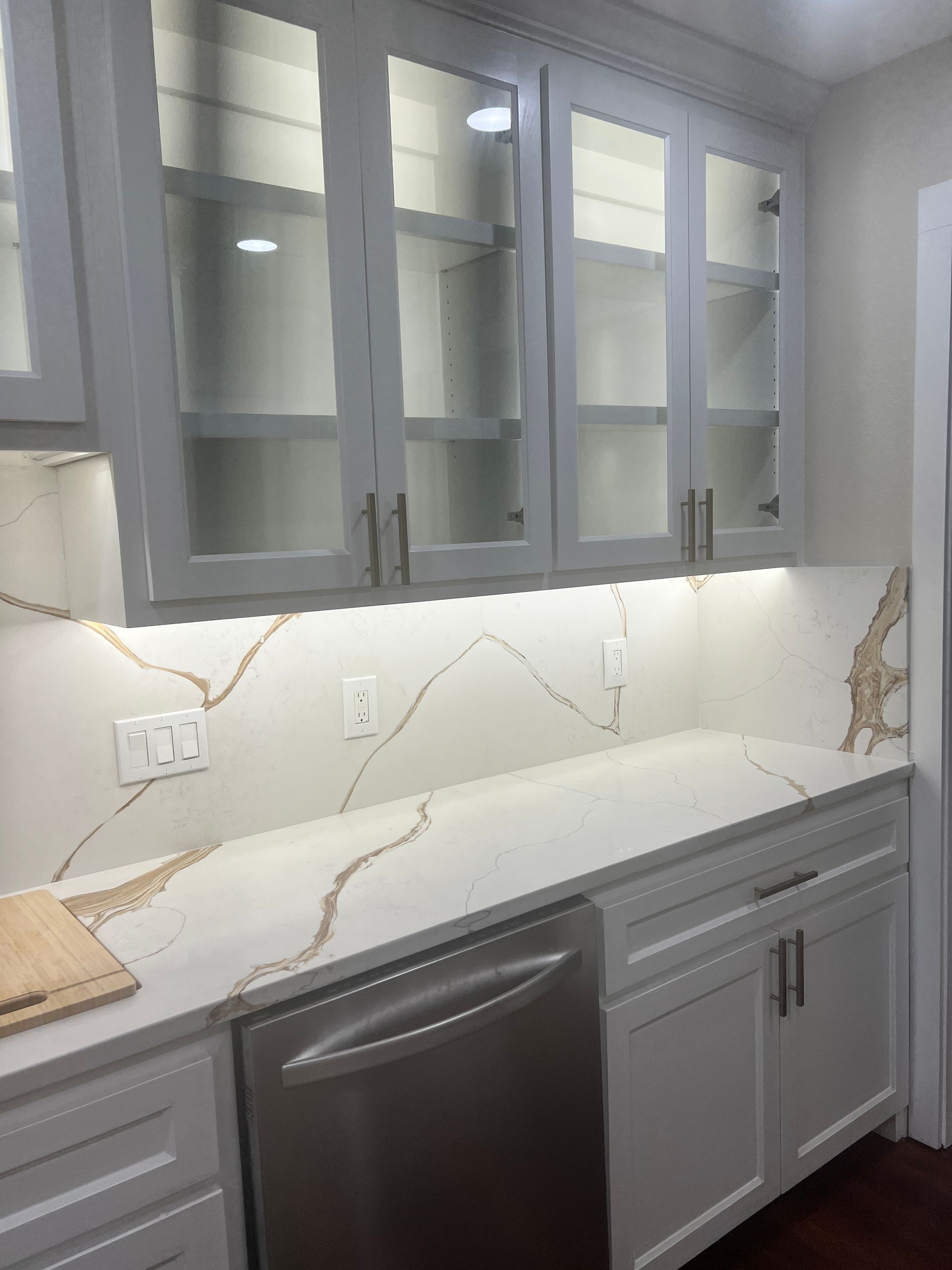 White kitchen with glass-front cabinets, gold veined countertops, and stainless steel dishwasher.