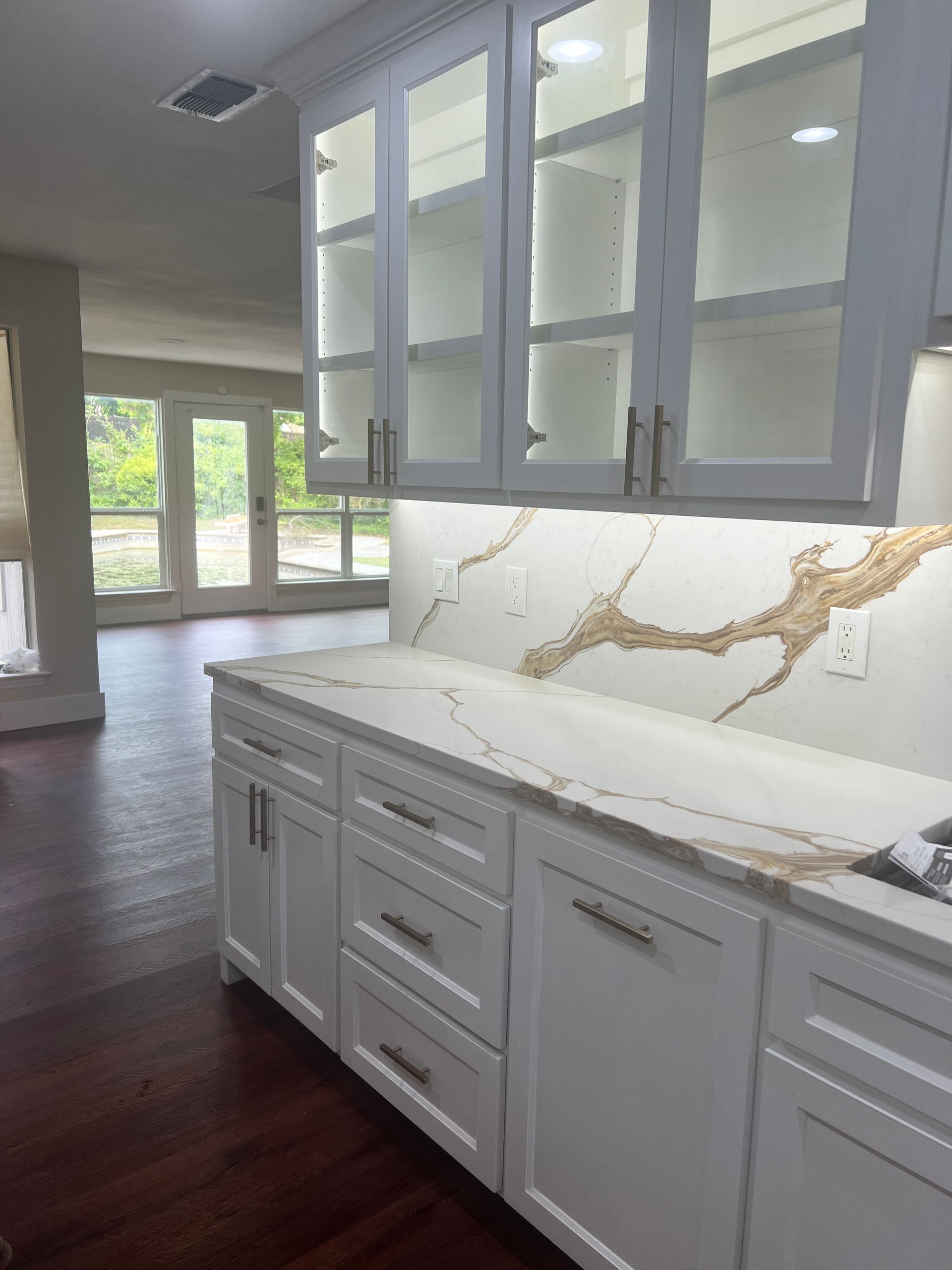 White kitchen cabinets with gold-veined countertops and glass-front uppers; view to living room.