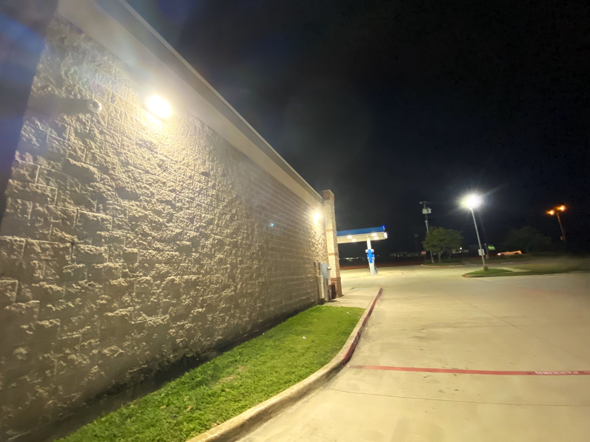 A brightly lit building's exterior wall at night with grass and a gas station visible.