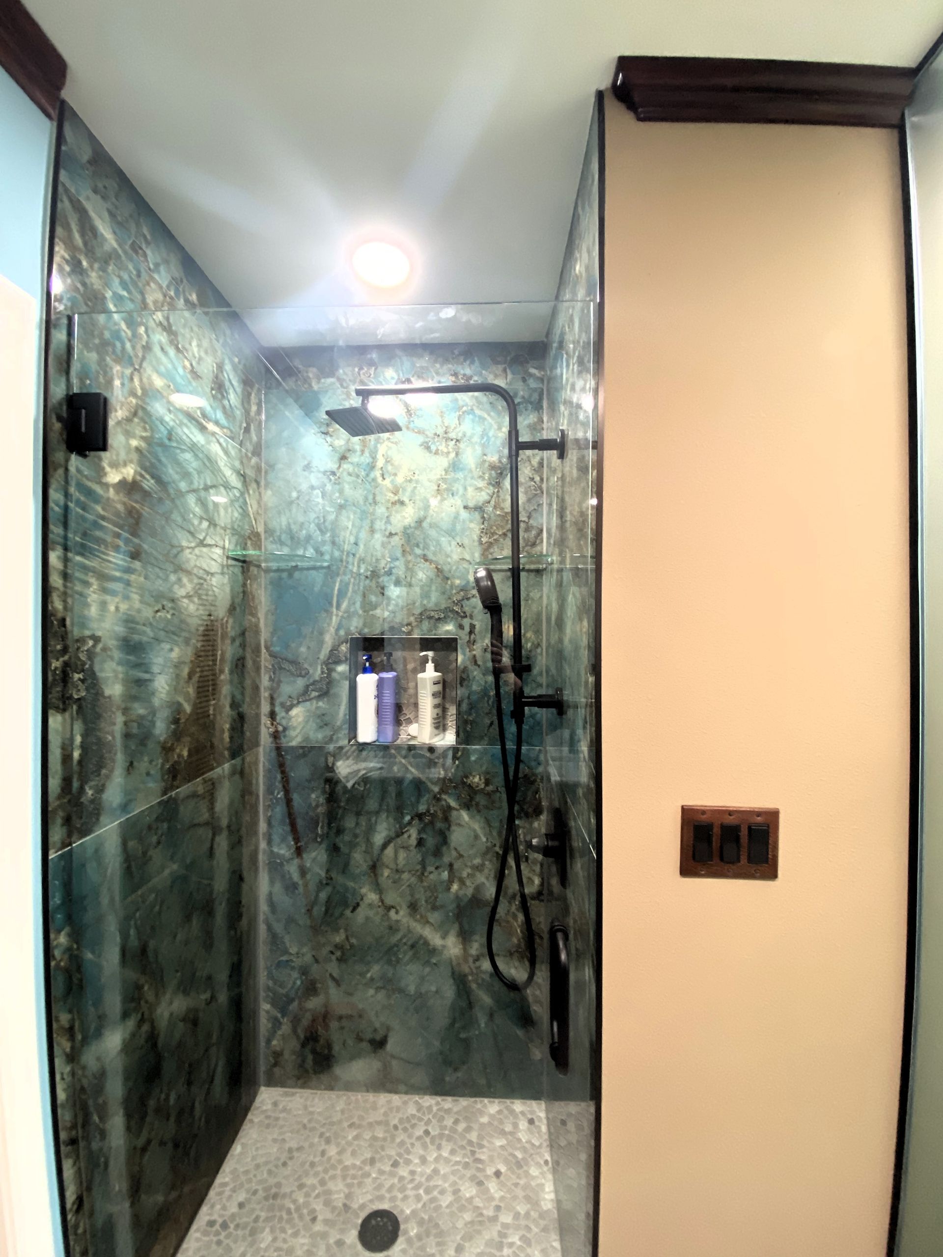 A modern shower with teal stone walls, glass door, dark fixtures, and a built-in shelf.