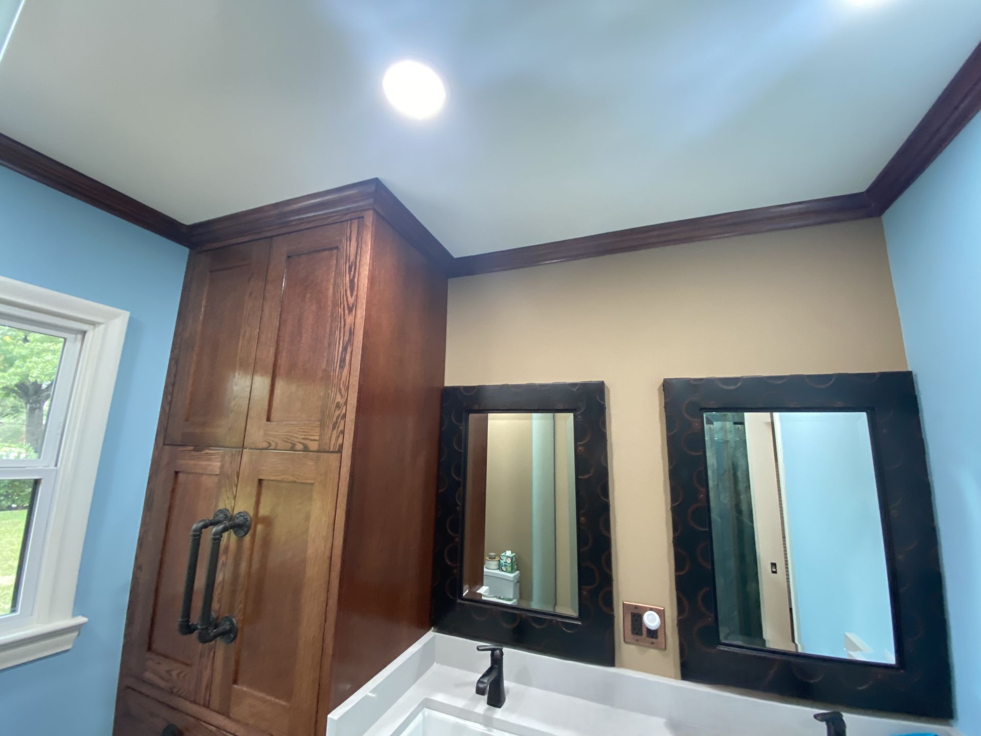 Bathroom with brown cabinets, tan and blue walls, black-framed mirrors, and a window.