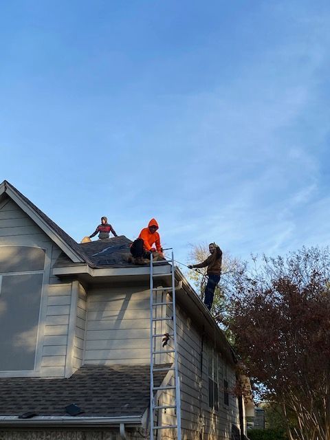 Three people on a roof: two on the shingles, one on the gutter, working on a house under a blue sky.