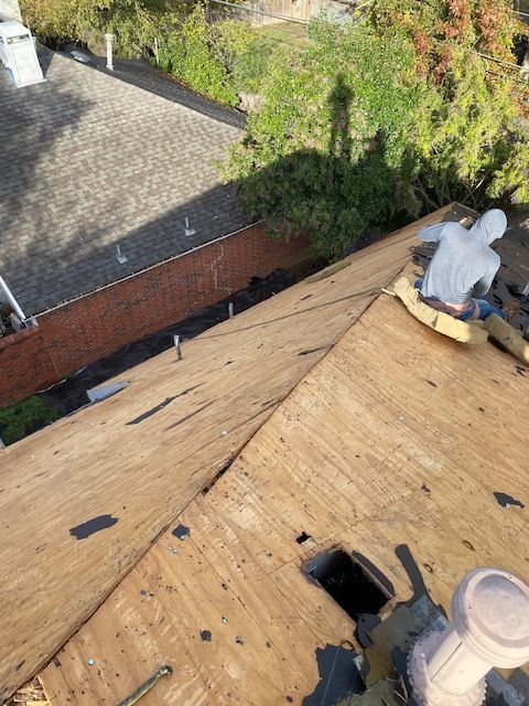 Roofer in white suit working on wood roof.  View from above, sunlit, surrounded by building and trees.