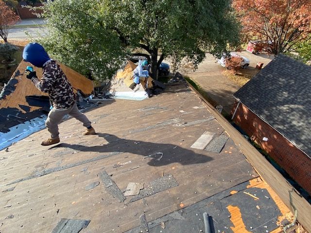 Roofers removing old shingles from a residential roof; one in camouflage throws debris.
