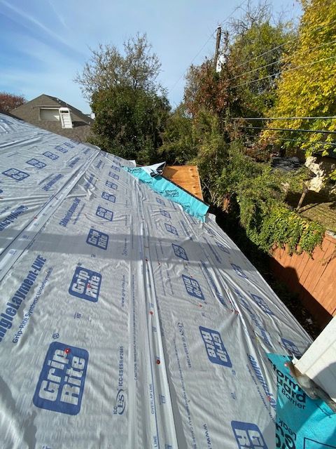 Roof covered with Grip-Rite underlayment; trees and brick wall visible in background.