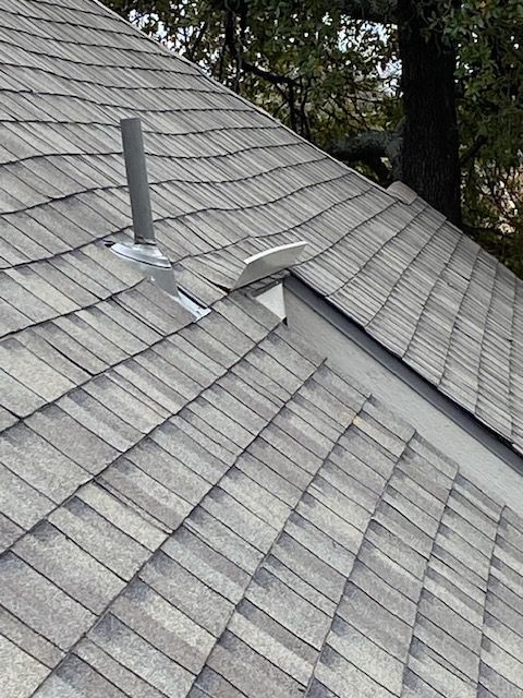 Gray shingle roof with a vent pipe and a triangular white vent against a tree backdrop.