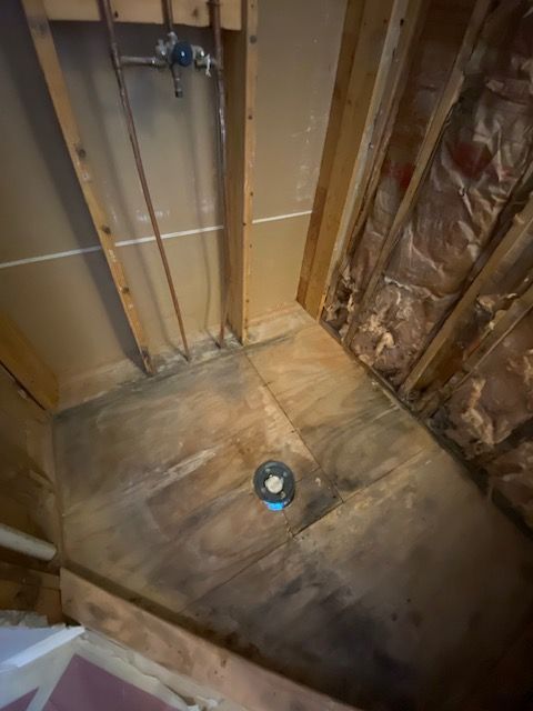 Wooden shower base with drain, copper pipes, and visible framing. Brown discoloration suggests water damage.