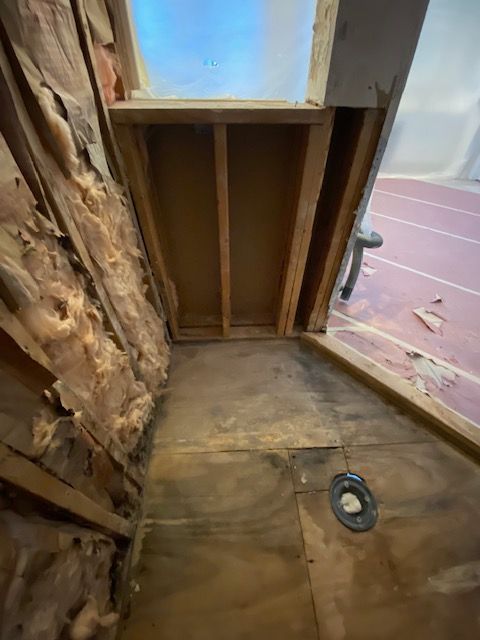 Interior view of a partially demolished shower stall. Exposed insulation, wooden framing, and a drain are visible.