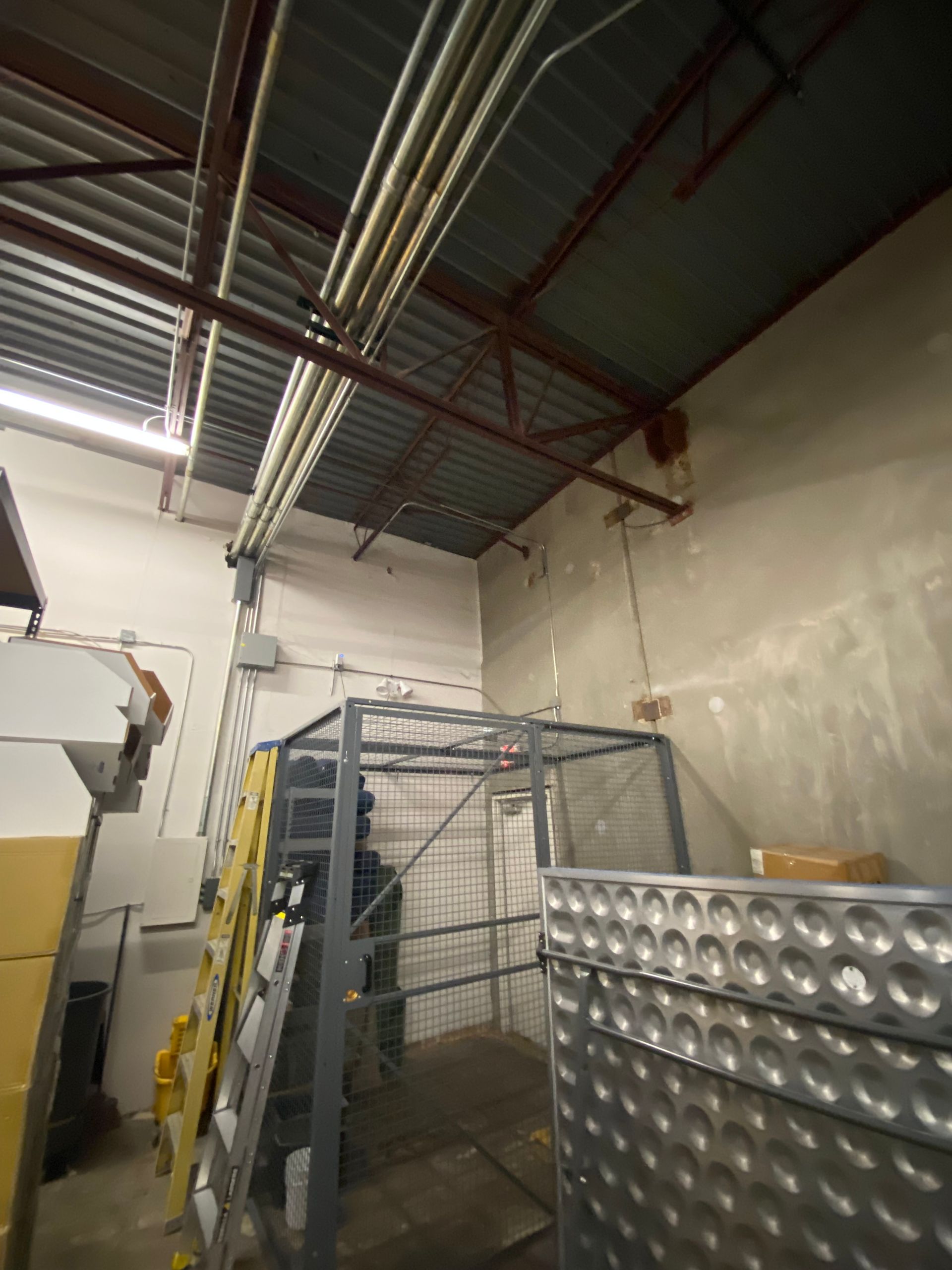 Metal cage and storage in a warehouse setting with overhead electrical conduits and high ceilings.