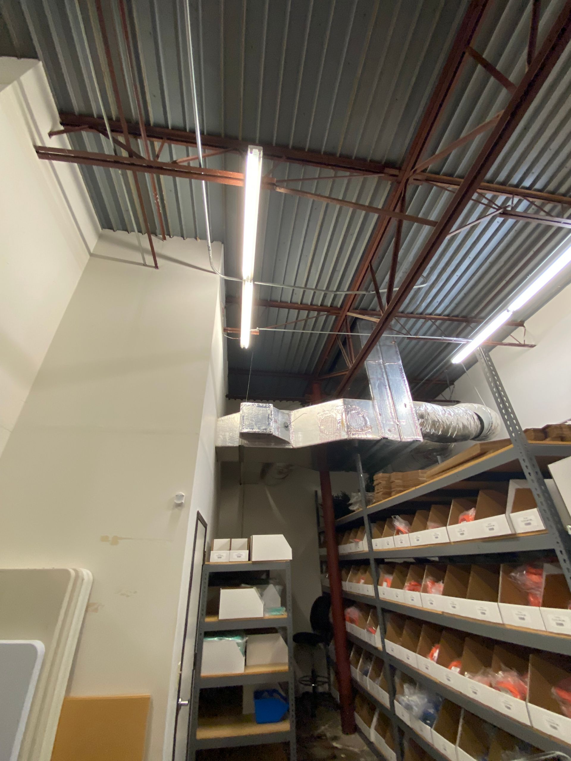 Warehouse interior with metal shelving, ventilation, and ceiling lights. A wall, shelving, and boxes are visible.