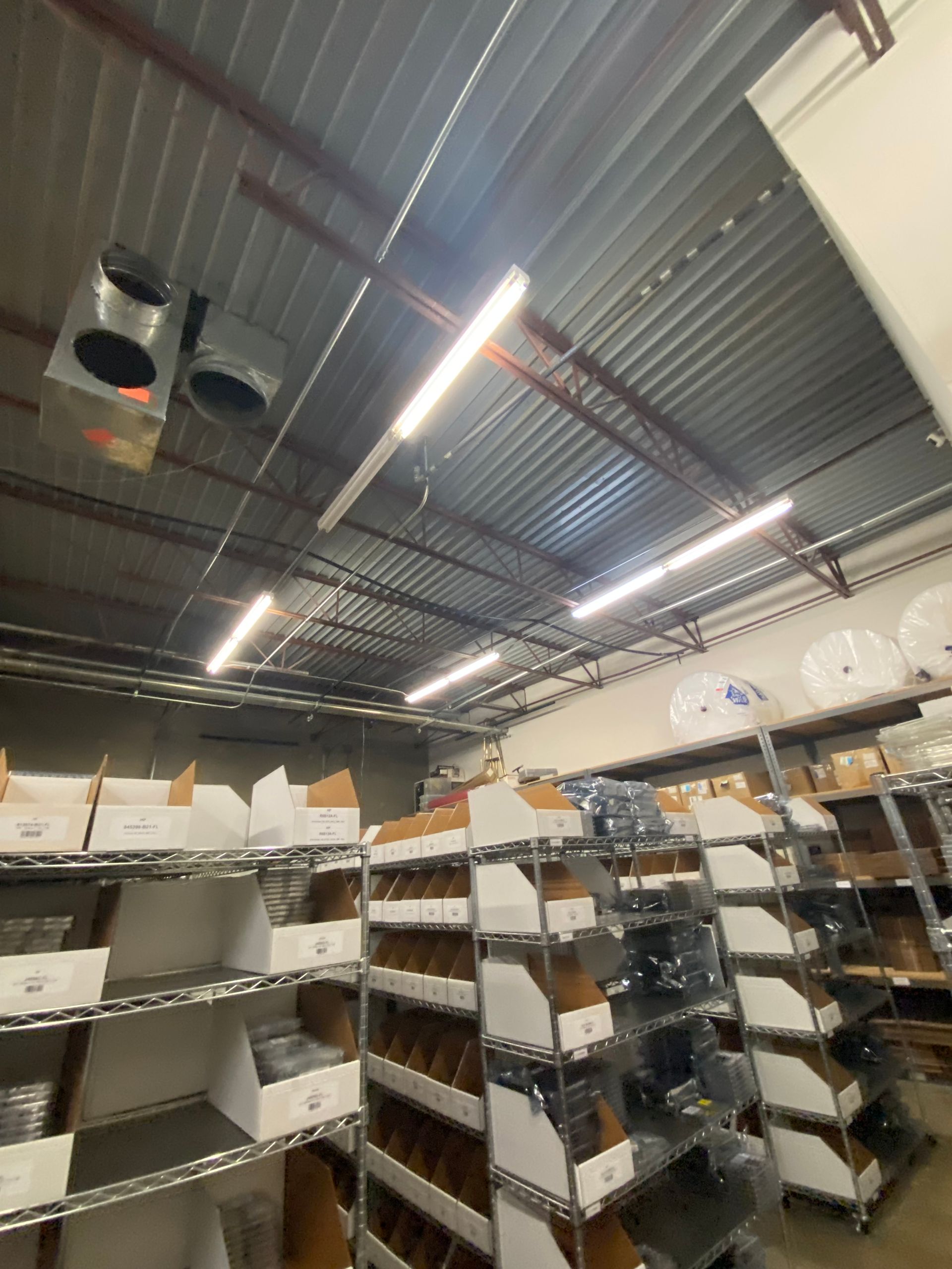 Warehouse interior with metal shelving and corrugated ceiling; boxes, parts, and overhead lighting.