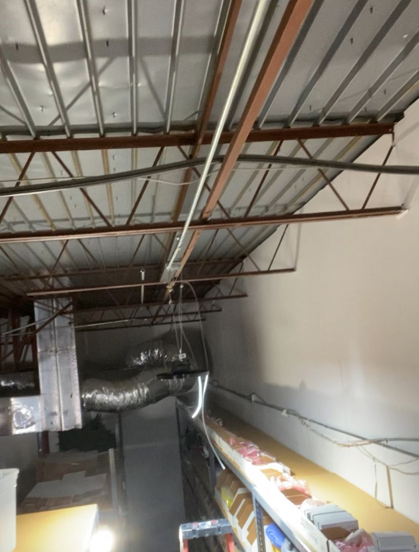 Warehouse ceiling with metal beams, ductwork, and a white wall.