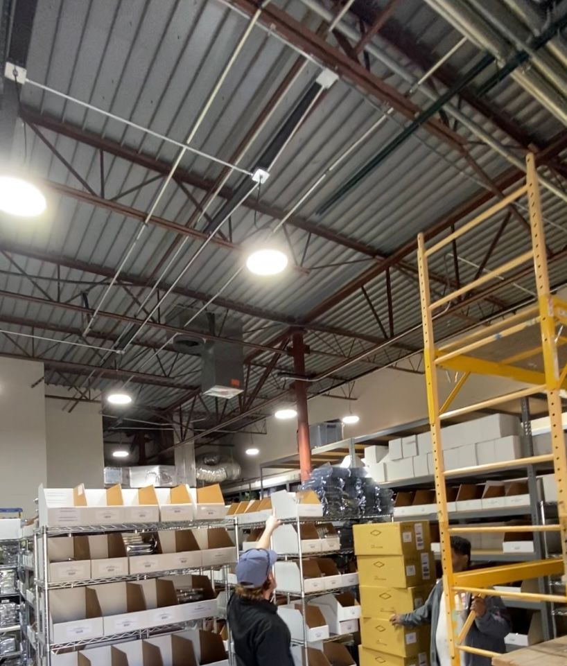 Warehouse interior: People, shelving, boxes, and overhead lighting. Steel beams and corrugated ceiling.