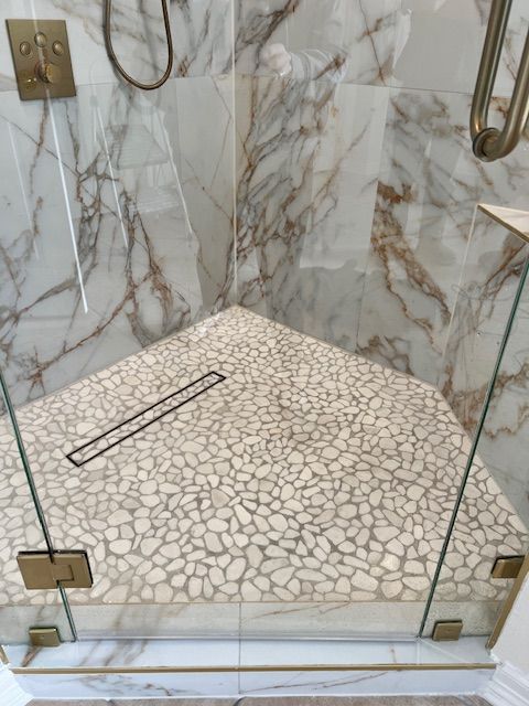 Shower with pebbled floor, marble walls, glass doors, and gold fixtures.
