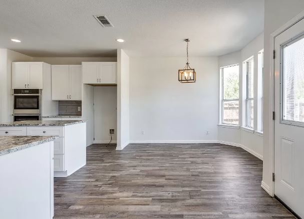 Empty dining area with white walls, hardwood floor, and a chandelier. Kitchen on left, windows on the right.