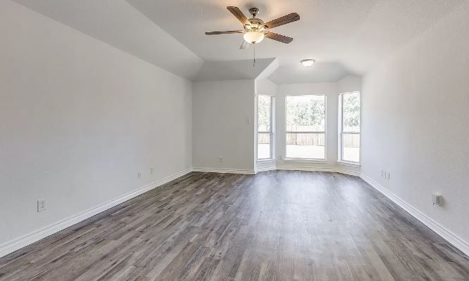 Empty room with gray wood-look floor, white walls, and a bay window, lit by a ceiling fan.