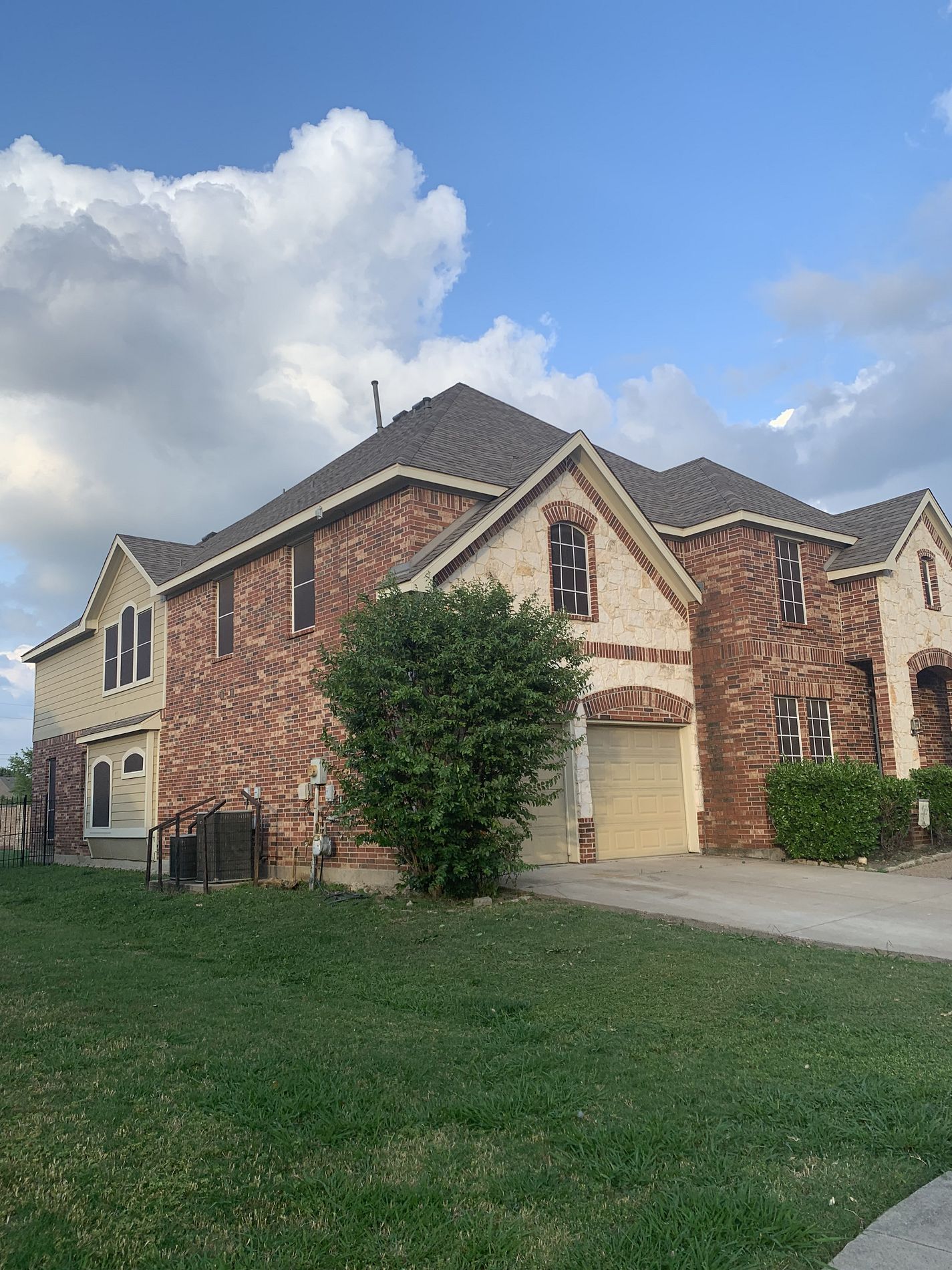 Two-story house with brick and beige siding, beige garage door, green grass, and cloudy sky.