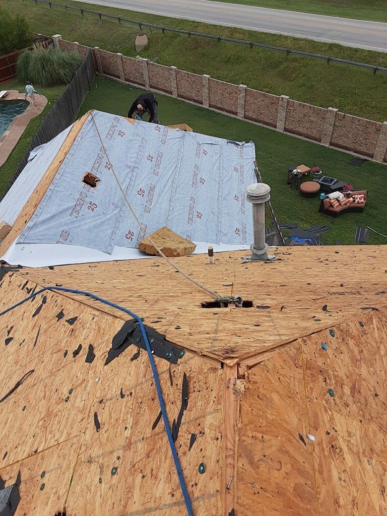 Roofer working on a house roof, partially covered with protective sheeting. Green lawn and brick fence in background.