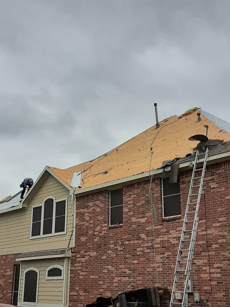 Roofer on a house removing shingles, exposing wood. Ladder leaning against red brick building under overcast sky.