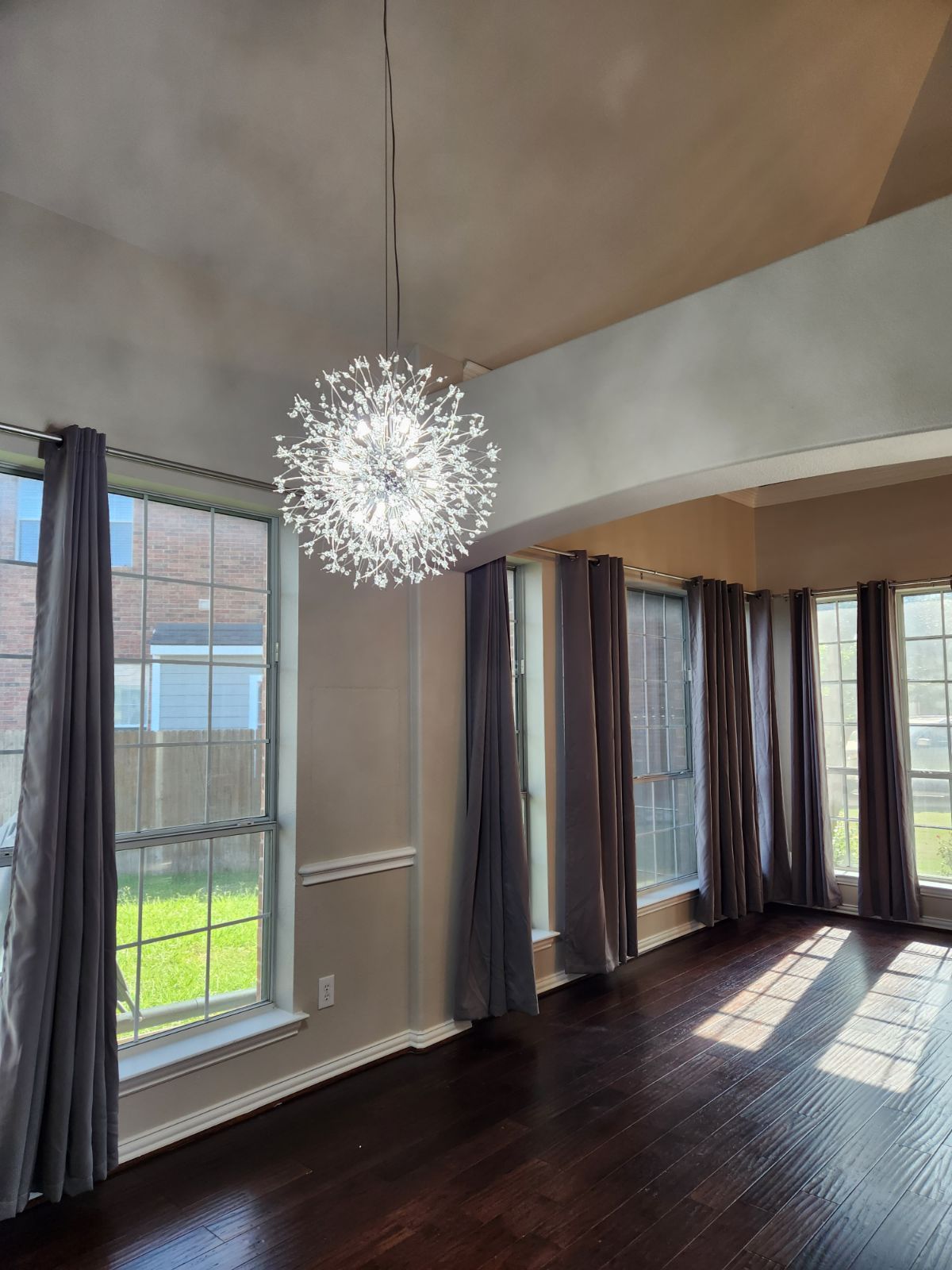 Room with dark wood floor, large windows, gray curtains, and a spherical chandelier.