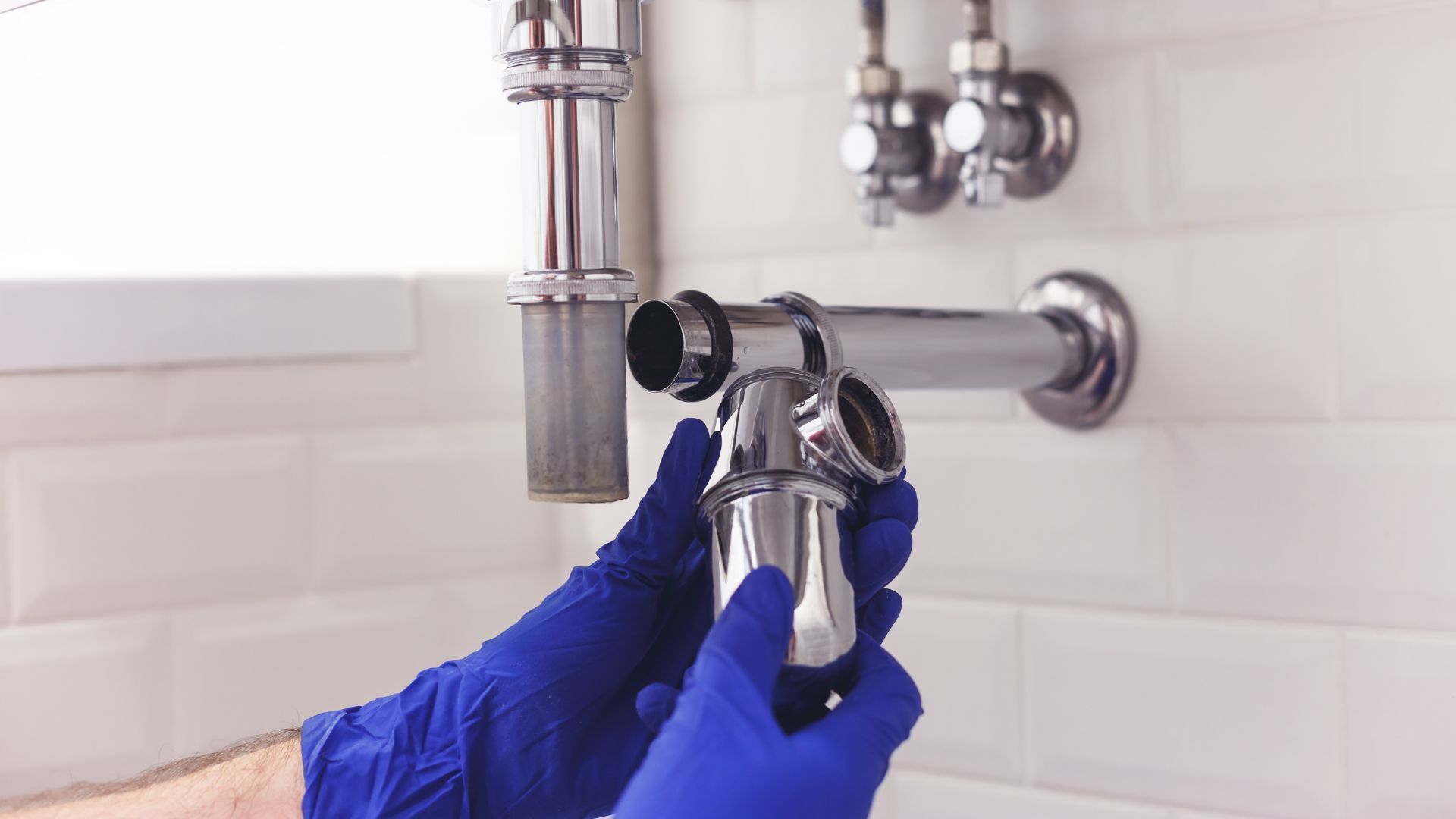Hands in blue gloves assembling a chrome bathroom P-trap pipe under a sink.