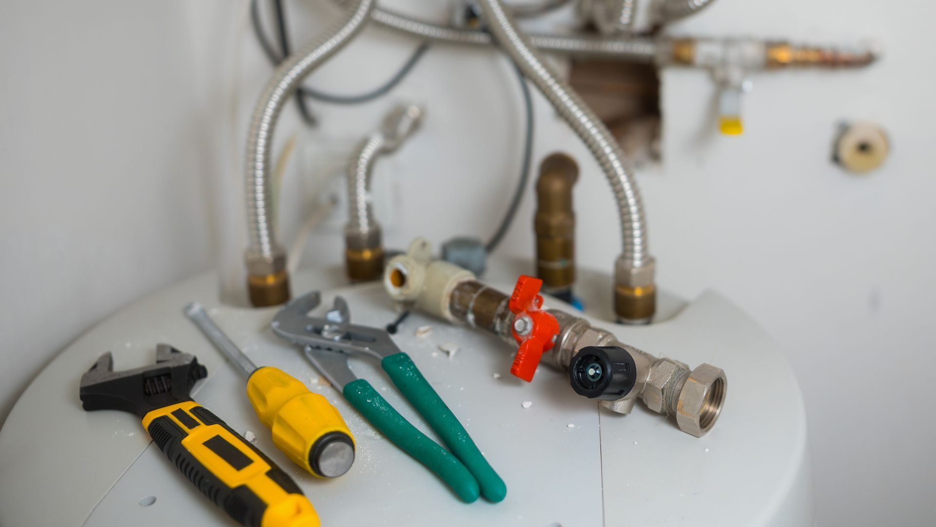 Plumbing tools, including an adjustable wrench, screwdriver, and pliers, resting on top of a water heater with pipe fittings.