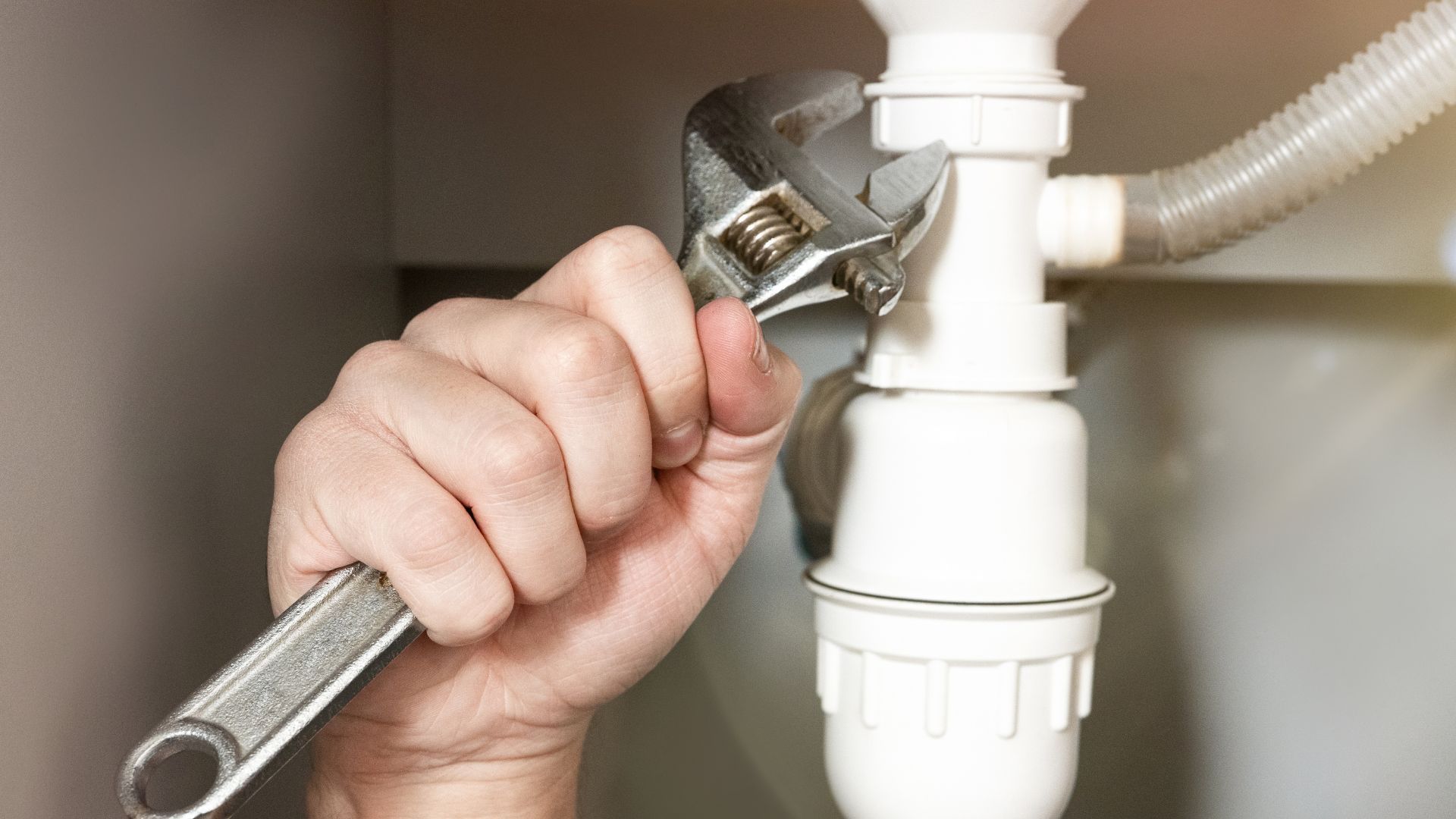 A person uses an adjustable wrench to tighten a white plastic plumbing pipe under a sink.