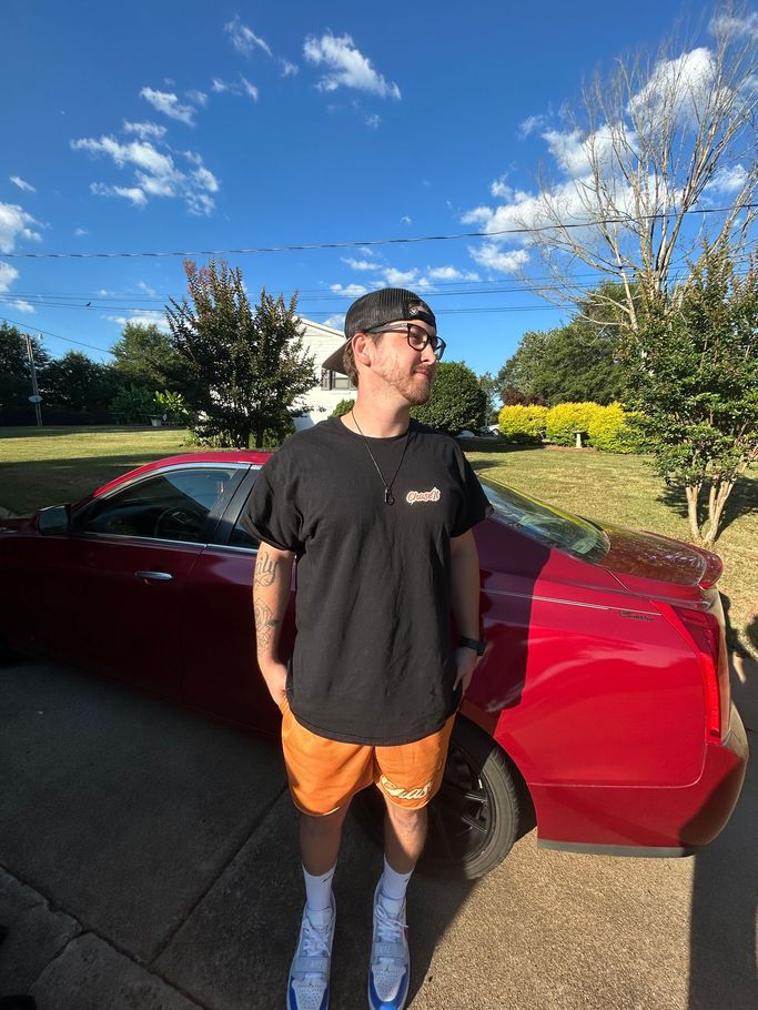 A man in a black shirt and orange shorts is standing in front of a red car.