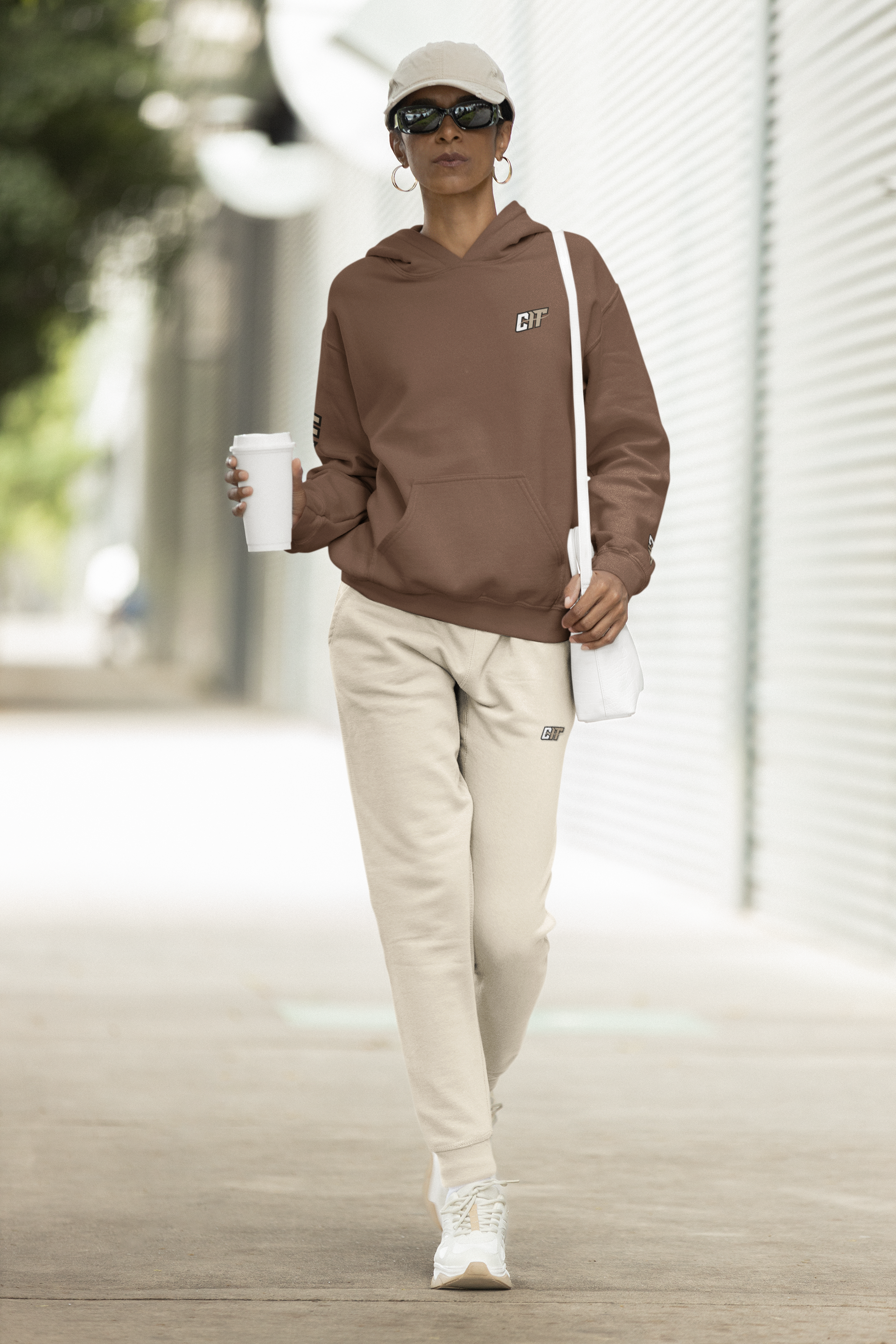 A woman wearing a brown hoodie and white sweatpants is walking down a sidewalk holding a cup of coffee.