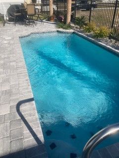 A large swimming pool is surrounded by a brick patio and a fence.