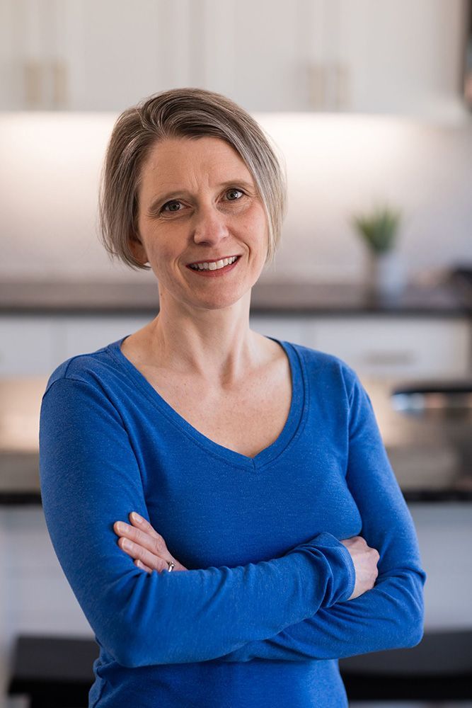 A woman in a blue shirt is standing in a kitchen with her arms crossed and smiling.