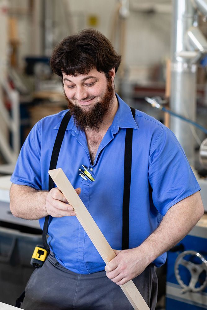 A man in a blue shirt and suspenders is holding a piece of wood.