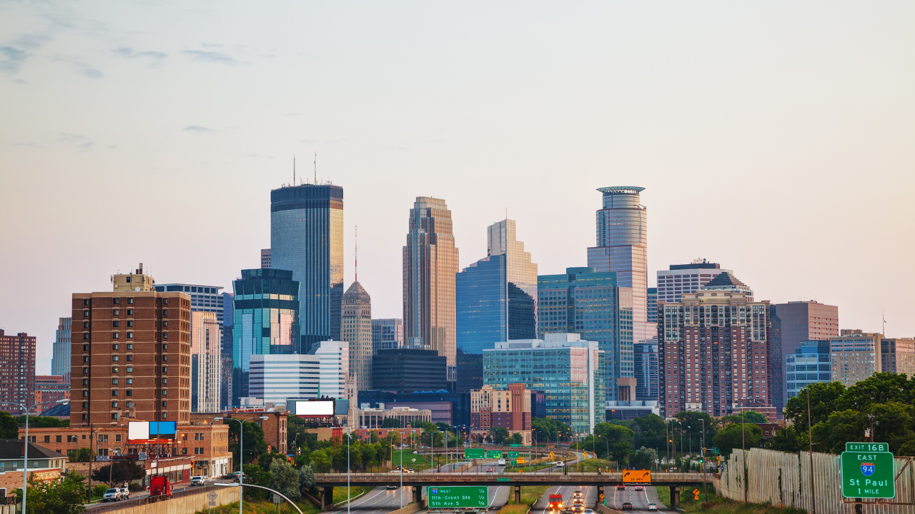 Skyline of Minneapolis, MN, with modern skyscrapers and highway in foreground at sunset.