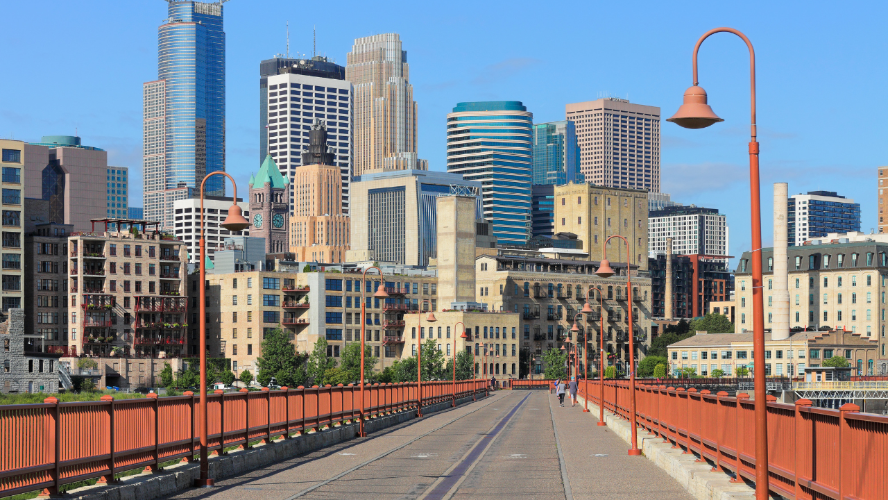 View of Minneapolis skyline from a bridge, with orange railing and light posts. Buildings vary in height and color.