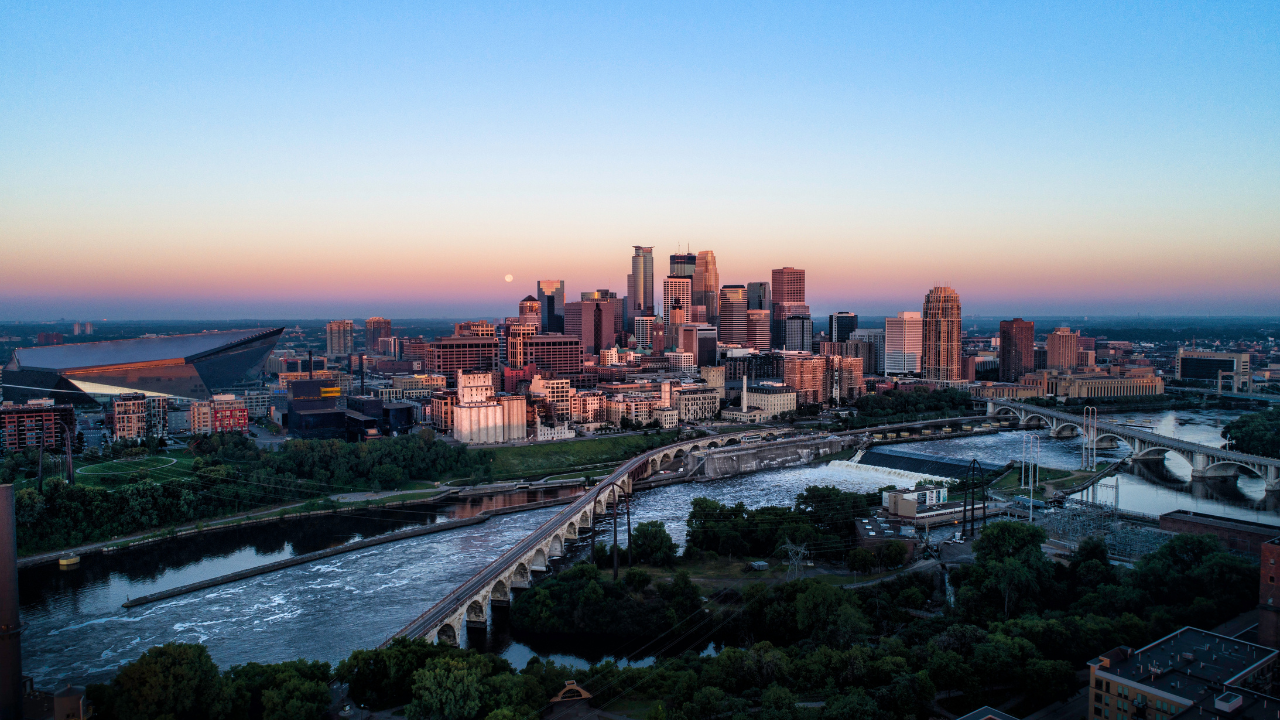 Minneapolis skyline at sunset, river and bridge in foreground, with blue and orange sky.